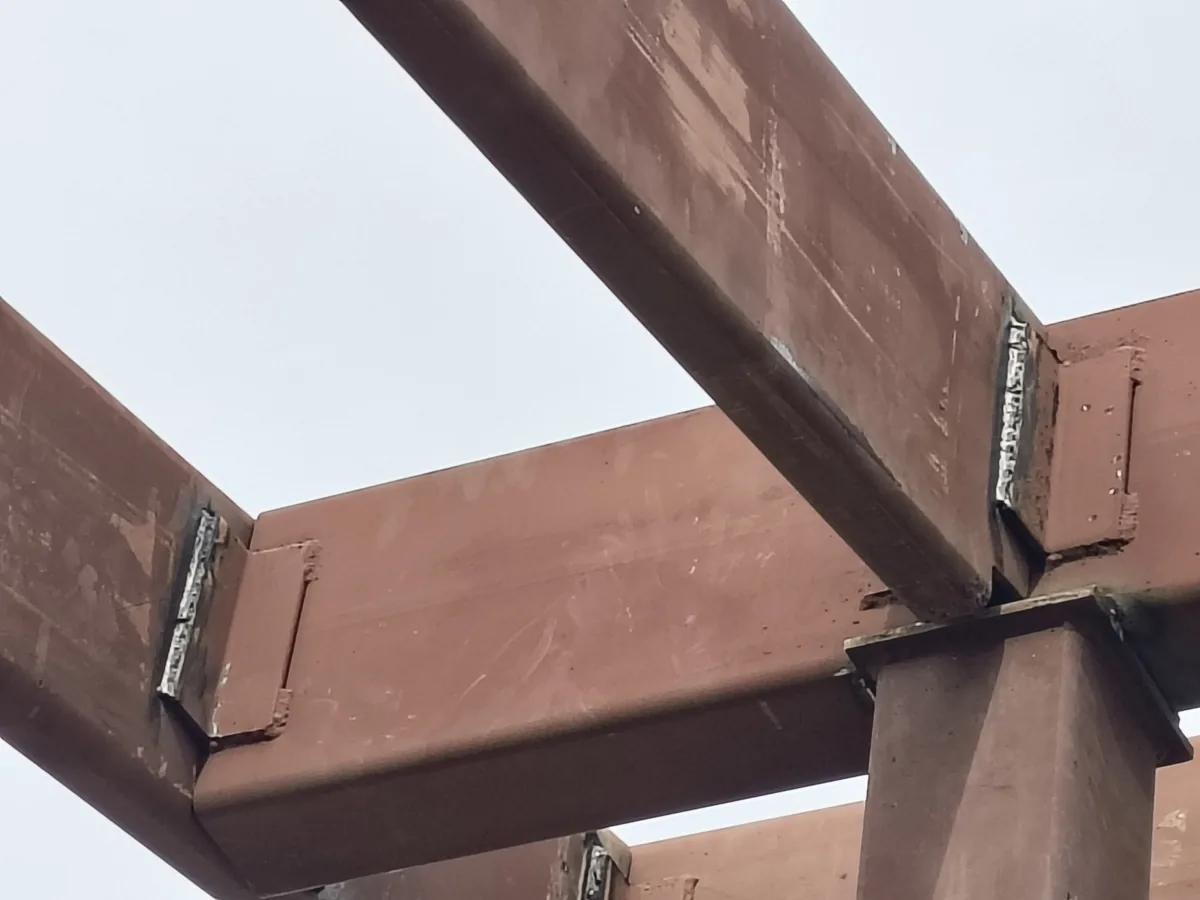 Close-up of a bolted and welded structural steel beam-to-column moment connection, primer-coated, viewed from below against an overcast sky.
