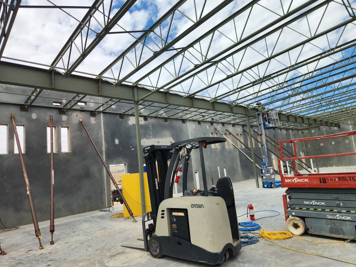 Interior of a steel-framed warehouse under construction, showing open-web bar joists and roof girders above a concrete slab with a forklift and scissor lift.