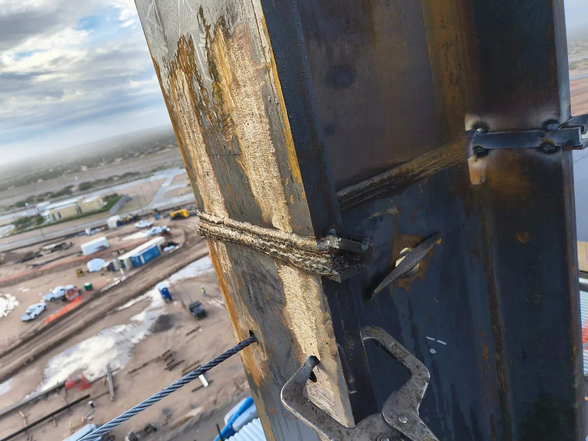 Close-up of a completed field weld on a steel column high above an active construction site, with the jobsite laydown yard visible far below.