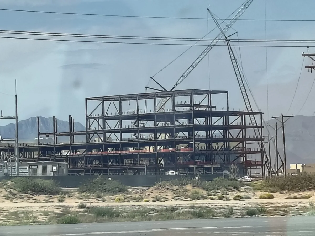 Multi-storey structural steel frame of a commercial building being erected by crane, seen from the roadside with the El Paso mountains behind it.